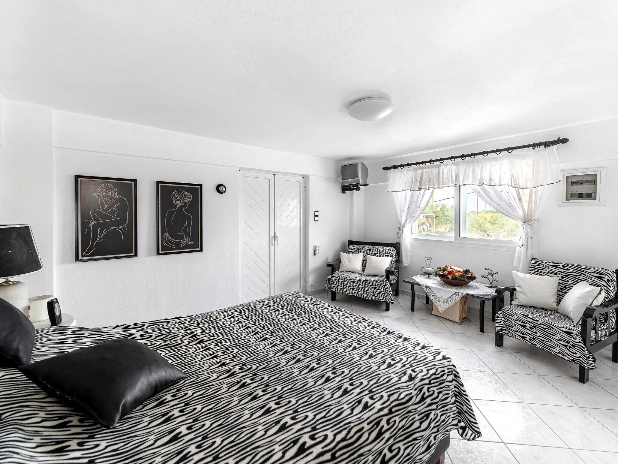 A bedroom with a zebra-patterned bedspread, two framed artworks, a wardrobe, a small table with a vase, and a window with curtains.