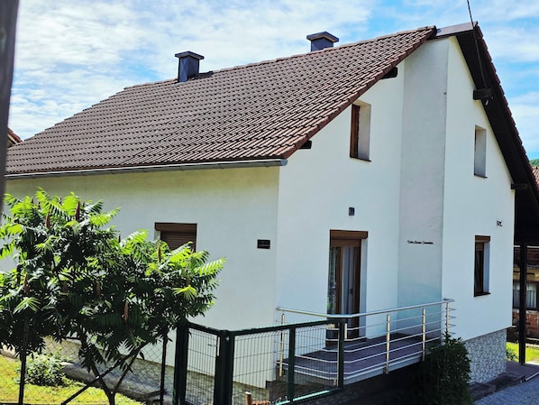 A two-story building with a tiled roof, a staircase leading to the entrance, and a small garden area with a tree.