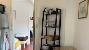 Une cuisine de taille réduite avec une table en bois, une étagère haute où sont rangés des bocaux et un réfrigérateur.