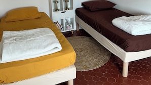 Une chambre avec deux lits, l’un recouvert d’une couvre-lit jaune et l’autre d’une couvre-lit marron, une table de chevet blanche avec une petite étagère, et une œuvre d’art encadrée accrochée au mur.