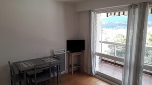 A room with a dining table and chairs, a television on a stand, and a large window with curtains. - Quiet studio, balcony with lake view, parking, near the spa, close to the center (aix les bains)
