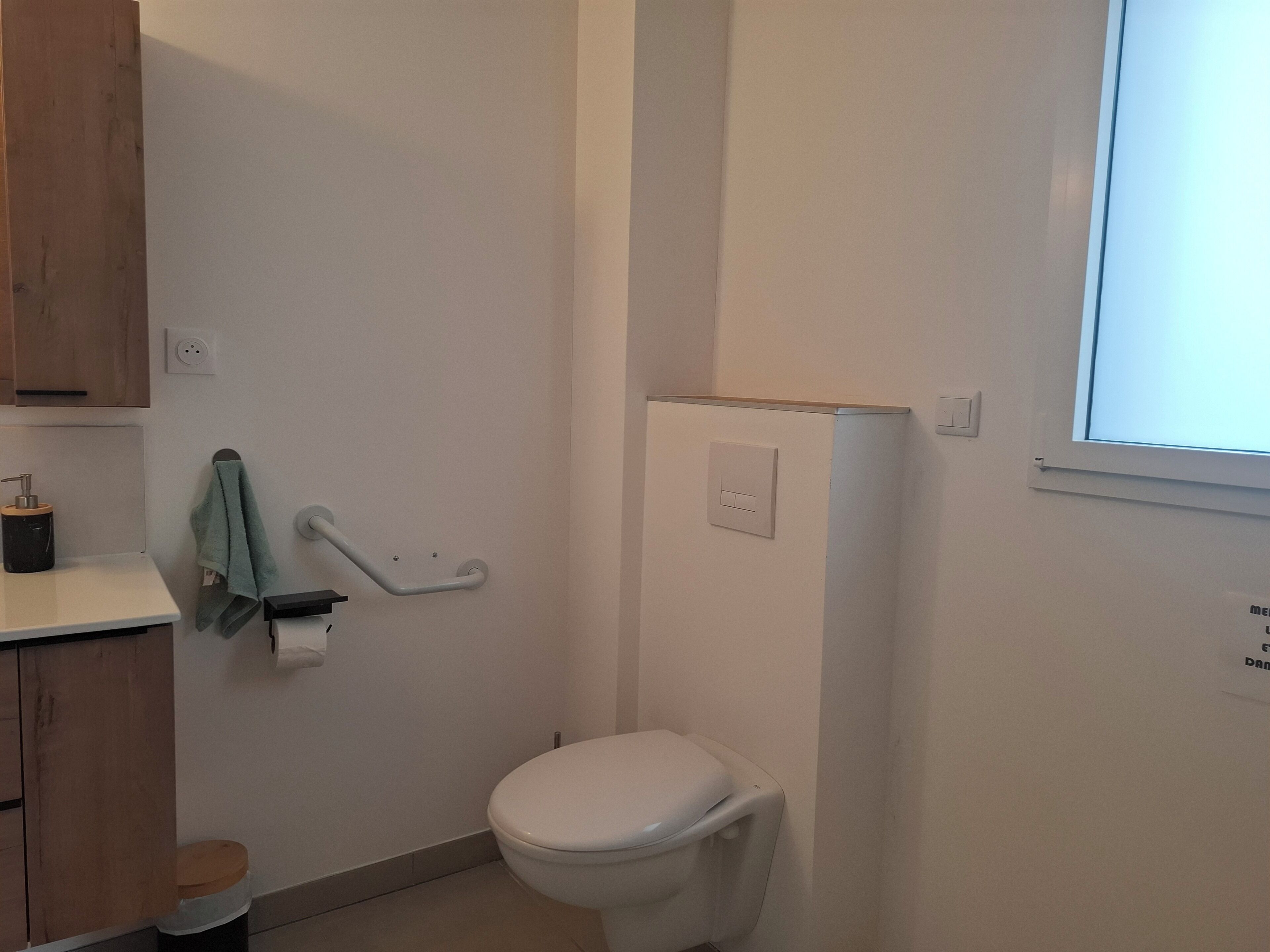 A small bathroom with a toilet, a handrail, a towel rack, and a window.