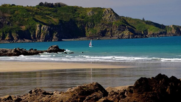 A coastal landscape with a sandy beach, rocky outcrops, and a sailboat on the water.