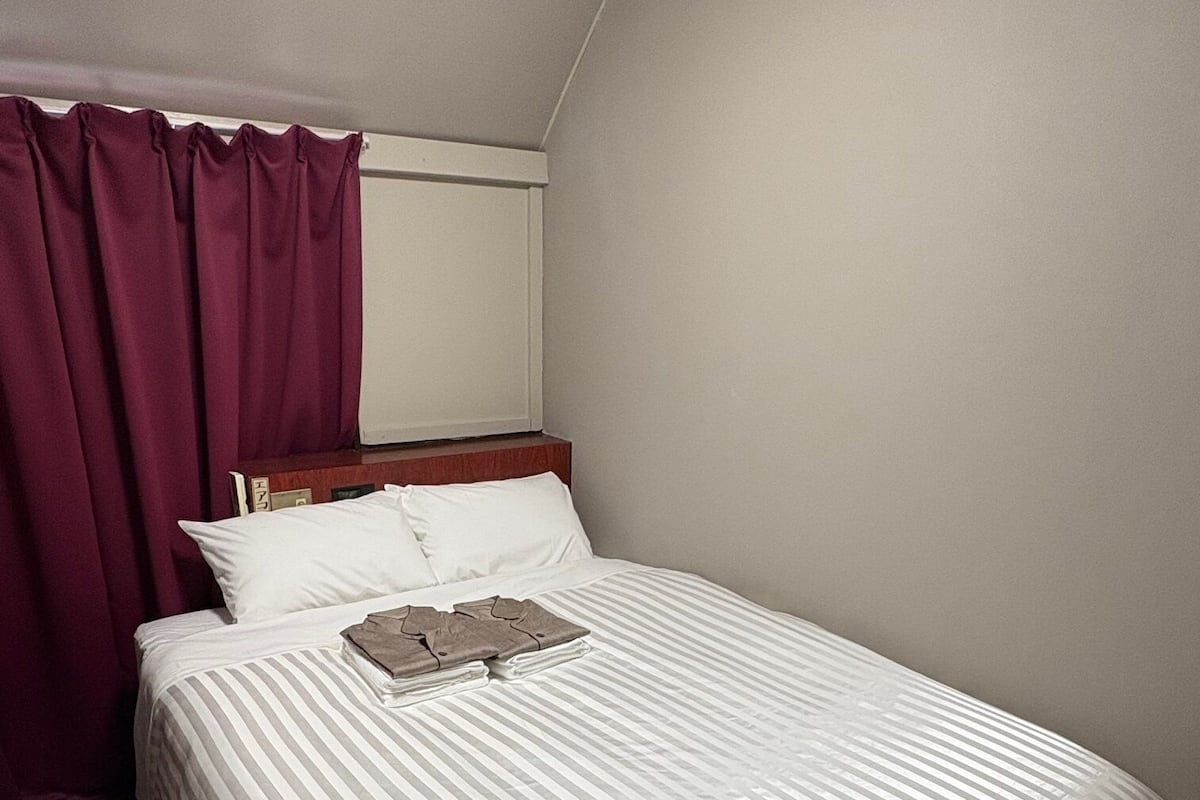 Une chambre d’hôtel avec un lit simple, une tête de lit en bois, une fenêtre avec des rideaux et un sol recouvert de moquette.