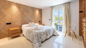 Un dormitorio moderno con una pared de madera en patrón espiga, una cama grande con ropa de cama estampada, una mesita de noche de madera, una silla transparente y vistas al balcón.