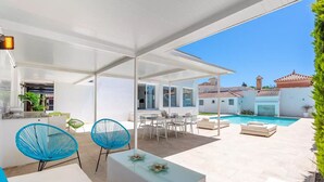 Un patio exterior moderno con piscina, muebles blancos y una zona de estar cubierta.