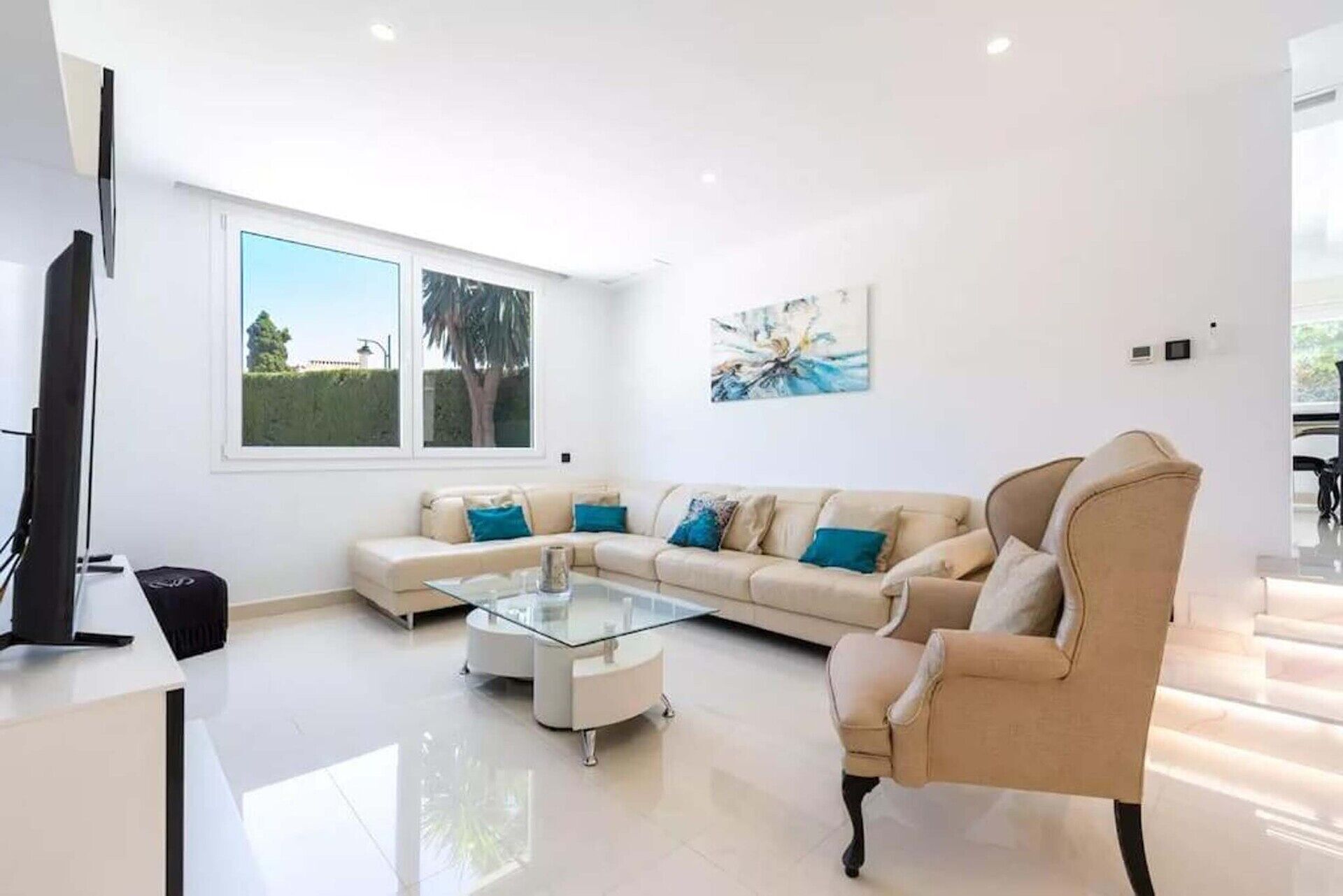 Ein modernes Wohnzimmer mit einem beigen Sofa, einem Glastisch und einem großen Fenster mit Blick auf Grünflächen.