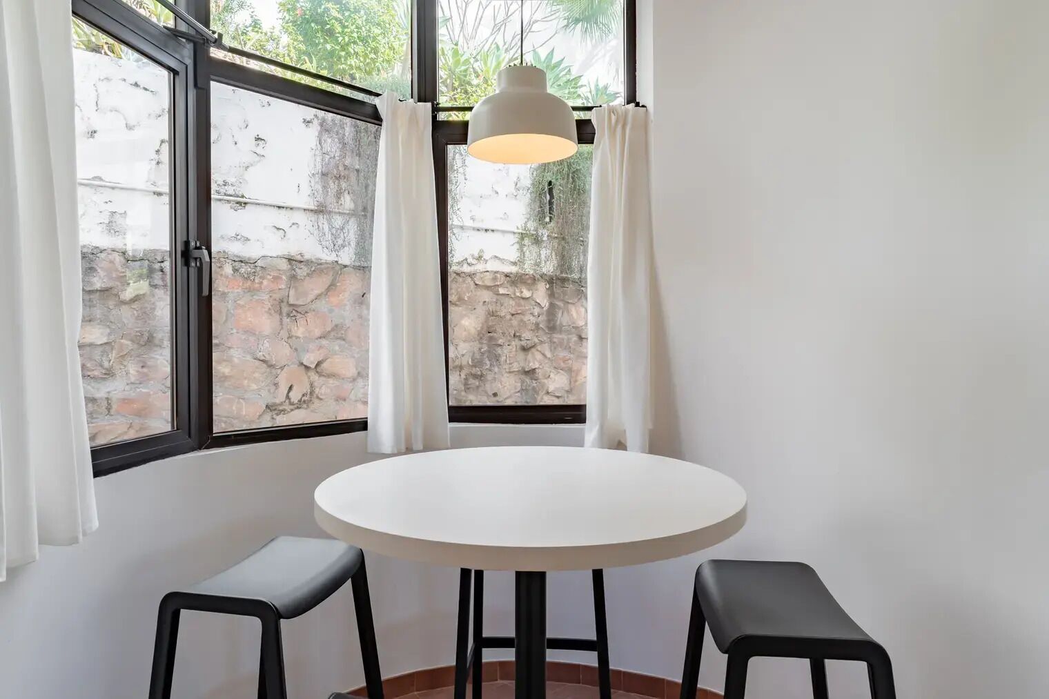 A round white table with two black stools, a hanging light fixture, and a view of a stone wall through a window with white curtains.