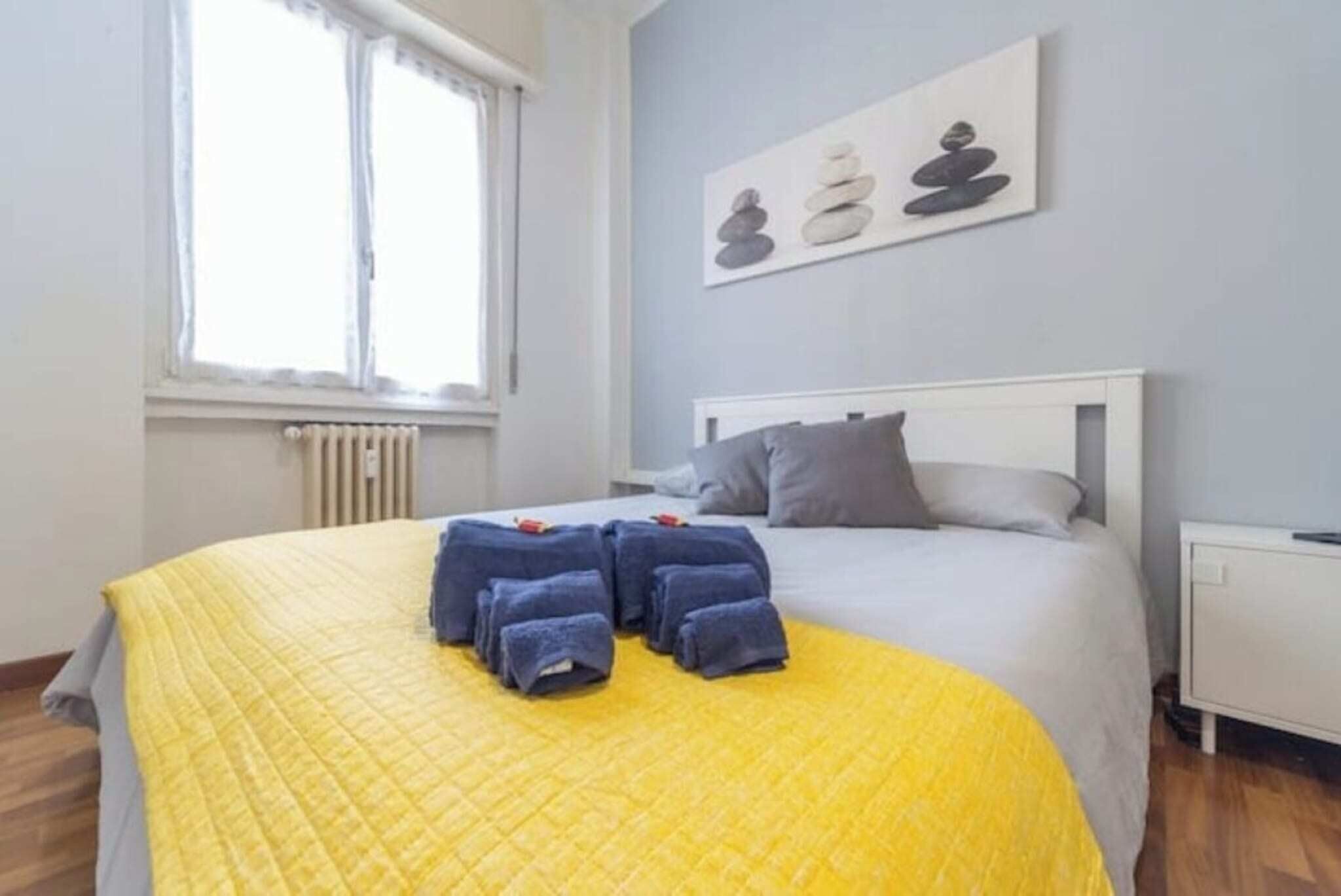 Een net opgemaakt bed met een gele sprei en een wit hoofdeinde. Handdoeken liggen op het bed, en aan de muur hangt een schilderij van gestapelde stenen.