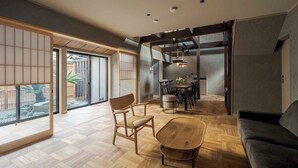 A spacious living area with a wooden floor, a sofa, a dining table, and a chair. There is a sliding door leading to a balcony with a view of the outside. - Waren Bettei Machiya House (Kyoto)
