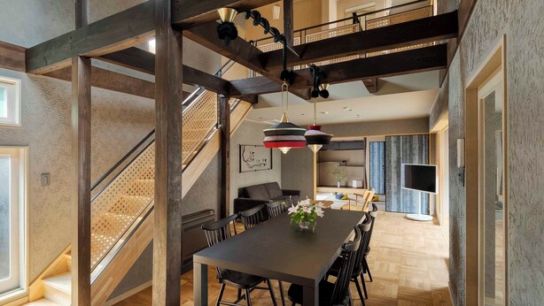 A spacious dining area with a long table and chairs, a staircase with a wooden railing, and a hanging pendant light. - Waren Bettei Machiya House (Kyoto)