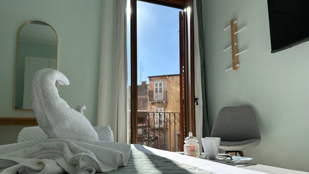 A hotel room with a bed, a chair, a table with a cup and a jar, and a view of buildings through the window. - Tropical style suite (Licata)