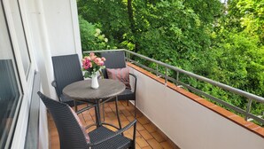 A balcony with two wicker chairs, a small round table with a vase of flowers, and a railing overlooking a lush green garden. - Family accommodation Moin Moin (Schilksee)