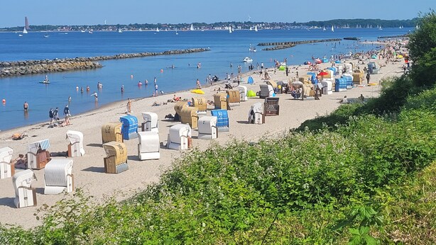A beach with numerous beach chairs and umbrellas, a rocky breakwater, and a distant view of boats and a lighthouse. - Family accommodation Moin Moin (Schilksee)