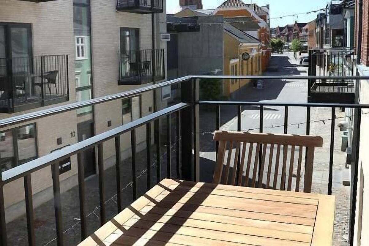 A balcony with a wooden table and chairs, overlooking a street with buildings and a church spire.