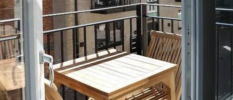 Un balcon avec une table et des chaises en bois, une porte vitrée, et un bâtiment aux murs de briques et aux balcons en arrière-plan.