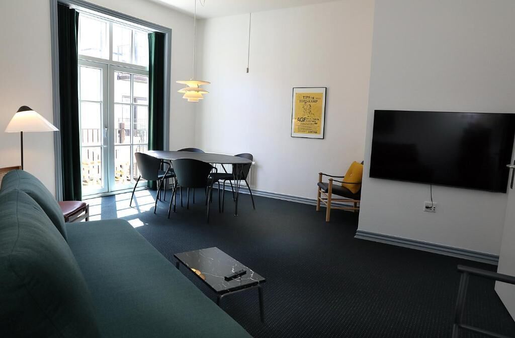 A modern living room with a sofa, a coffee table, a TV, a dining area with chairs, and a large window.