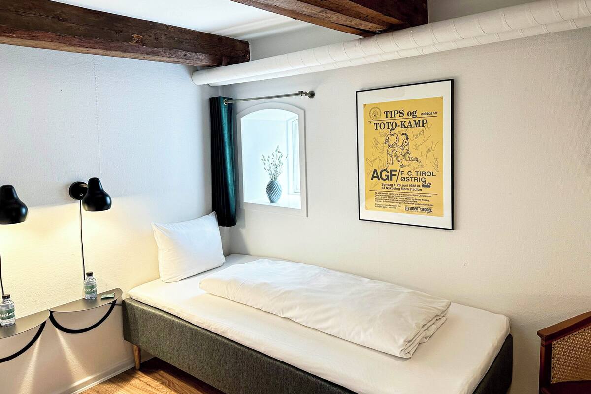 A small, neatly made bed in a room with a wooden ceiling beam, a framed poster on the wall, and a window with a curtain.