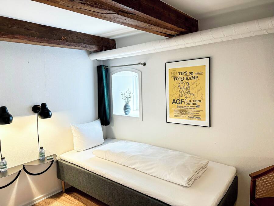 Ein kleines, ordentlich bezogenes Bett in einem Zimmer mit Holzbalkendecke, einem gerahmten Poster an der Wand und einem Fenster mit Vorhang.