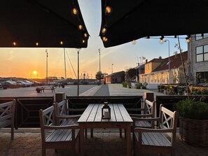 Un espace extérieur aménagé avec des bancs et des tables en bois, un coucher de soleil en arrière-plan et un parking avec des voitures.