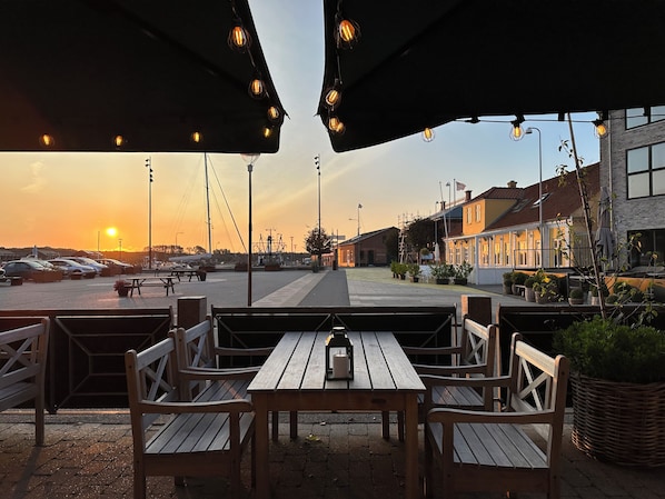 An outdoor seating area with wooden benches and tables, a sunset in the background, and a parking lot with cars. - Hotel Steenbergs (Nykøbing Mors)