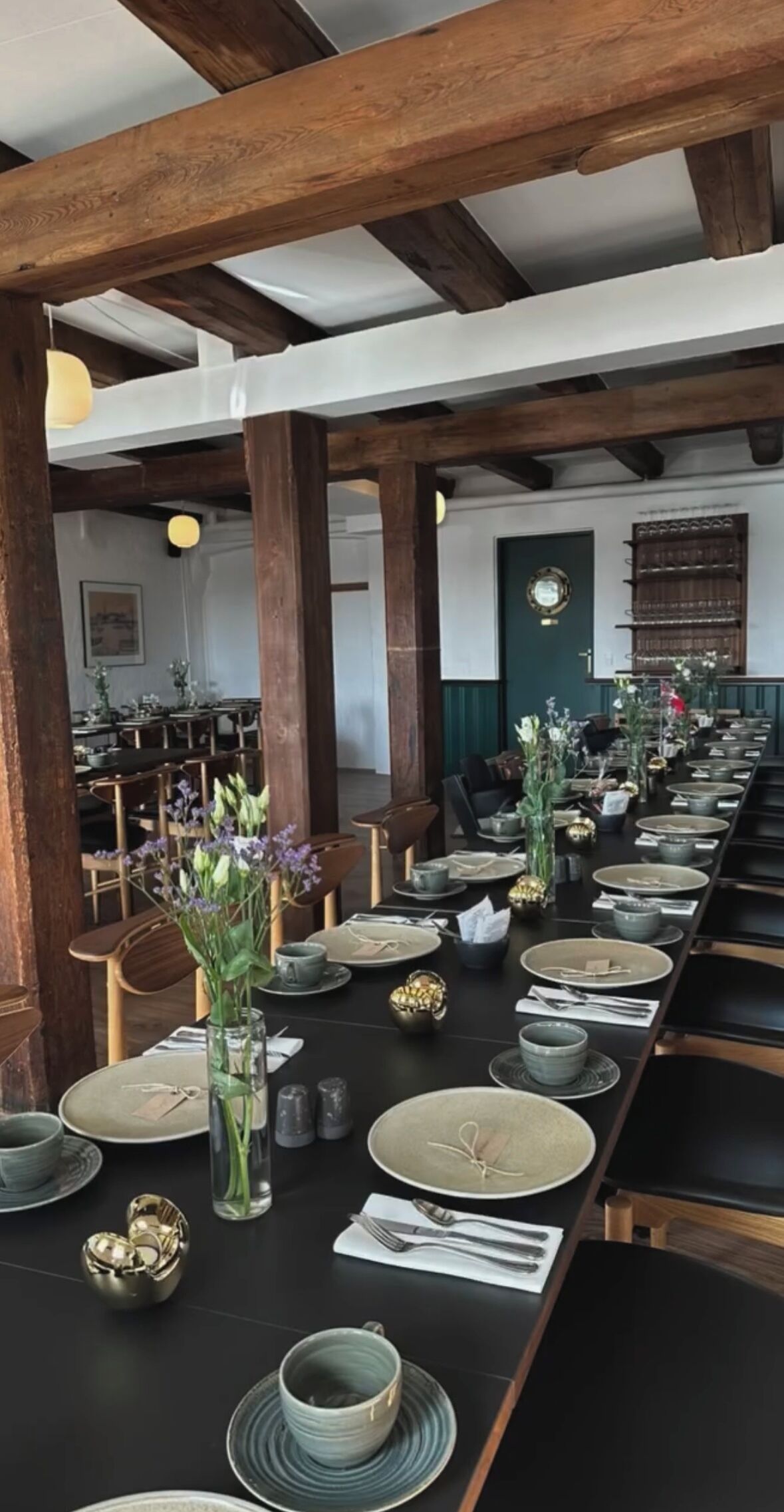 A long dining table set with plates, bowls, and cutlery, surrounded by wooden chairs and a dark tablecloth.