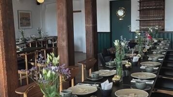 A long dining table set with plates, bowls, and cutlery, surrounded by wooden chairs and a dark tablecloth.