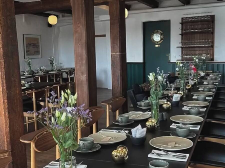 A long dining table set with plates, bowls, and cutlery, surrounded by wooden chairs and a dark tablecloth.