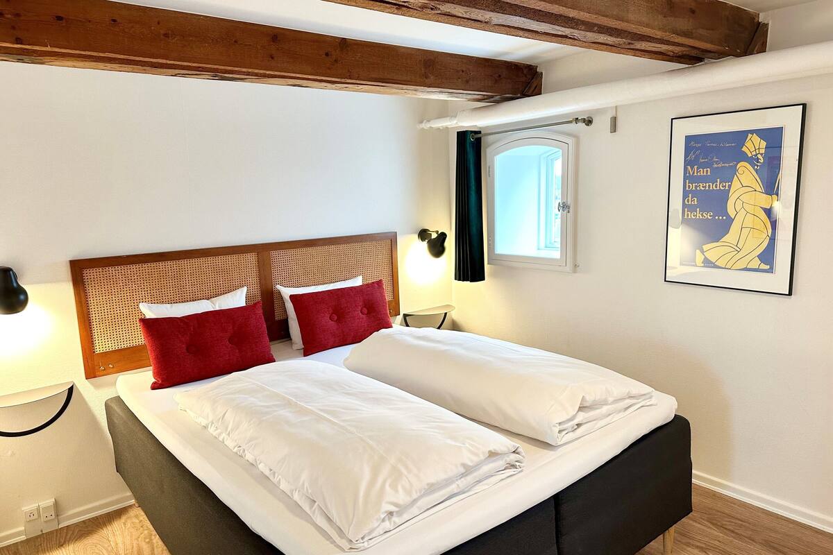 A hotel room with a bed, wooden headboard, red pillows, white bedding, a window with curtains, a wall-mounted lamp, and a framed poster.