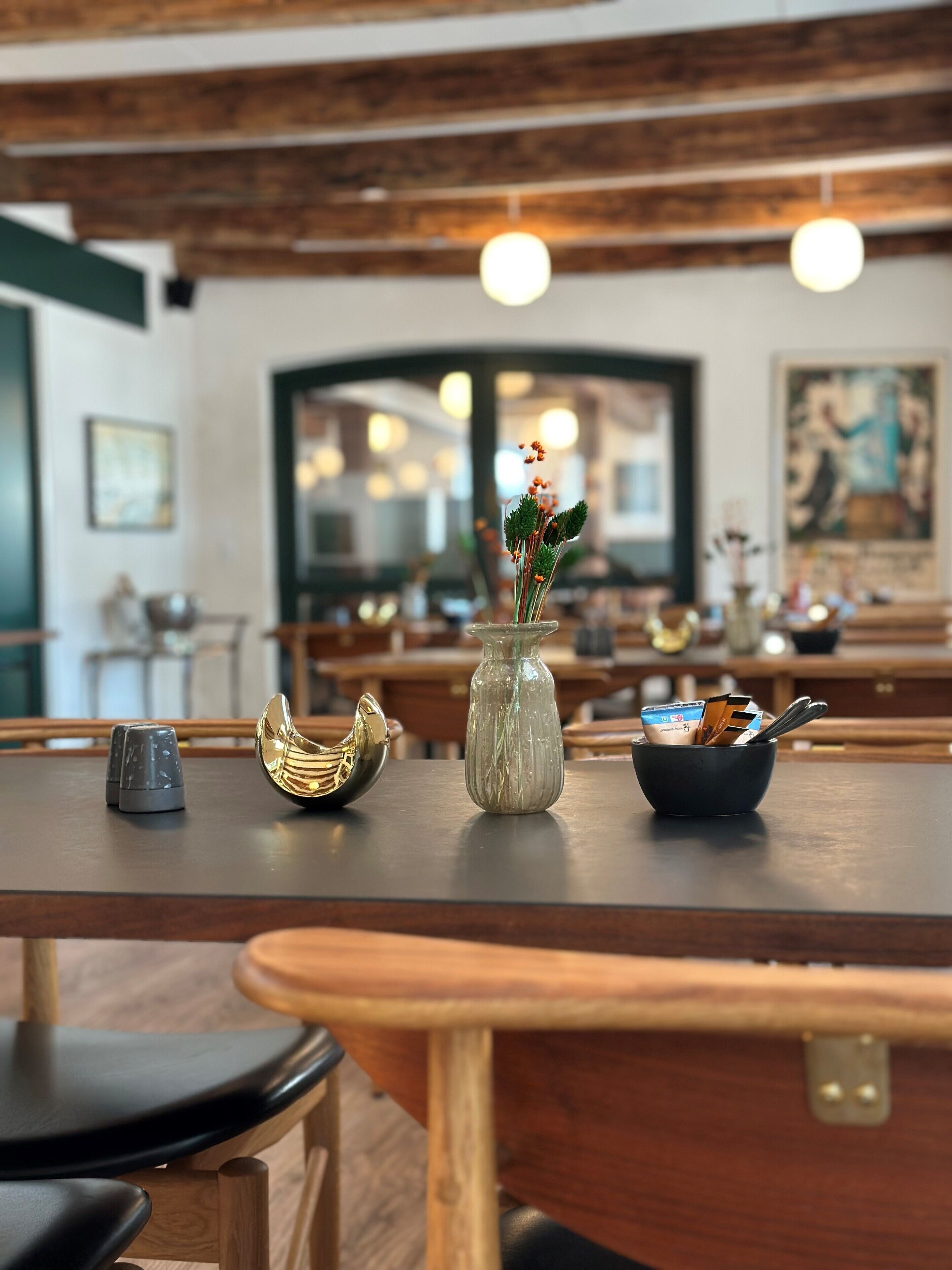 Interior de restaurante con mesas de madera, un jarrón con flores y un bol con cubiertos.