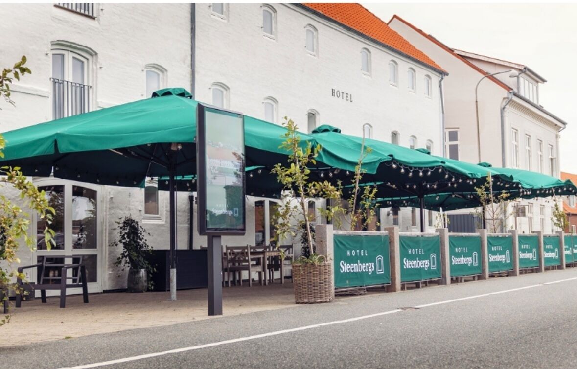 A hotel with outdoor seating under green awnings, a signboard, and a row of identical signs along the sidewalk.