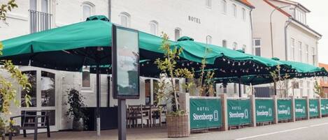 A hotel with outdoor seating under green awnings, a signboard, and a row of identical signs along the sidewalk.