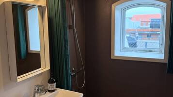 A bathroom with a sink, mirror, shower, and a window with a view of buildings.