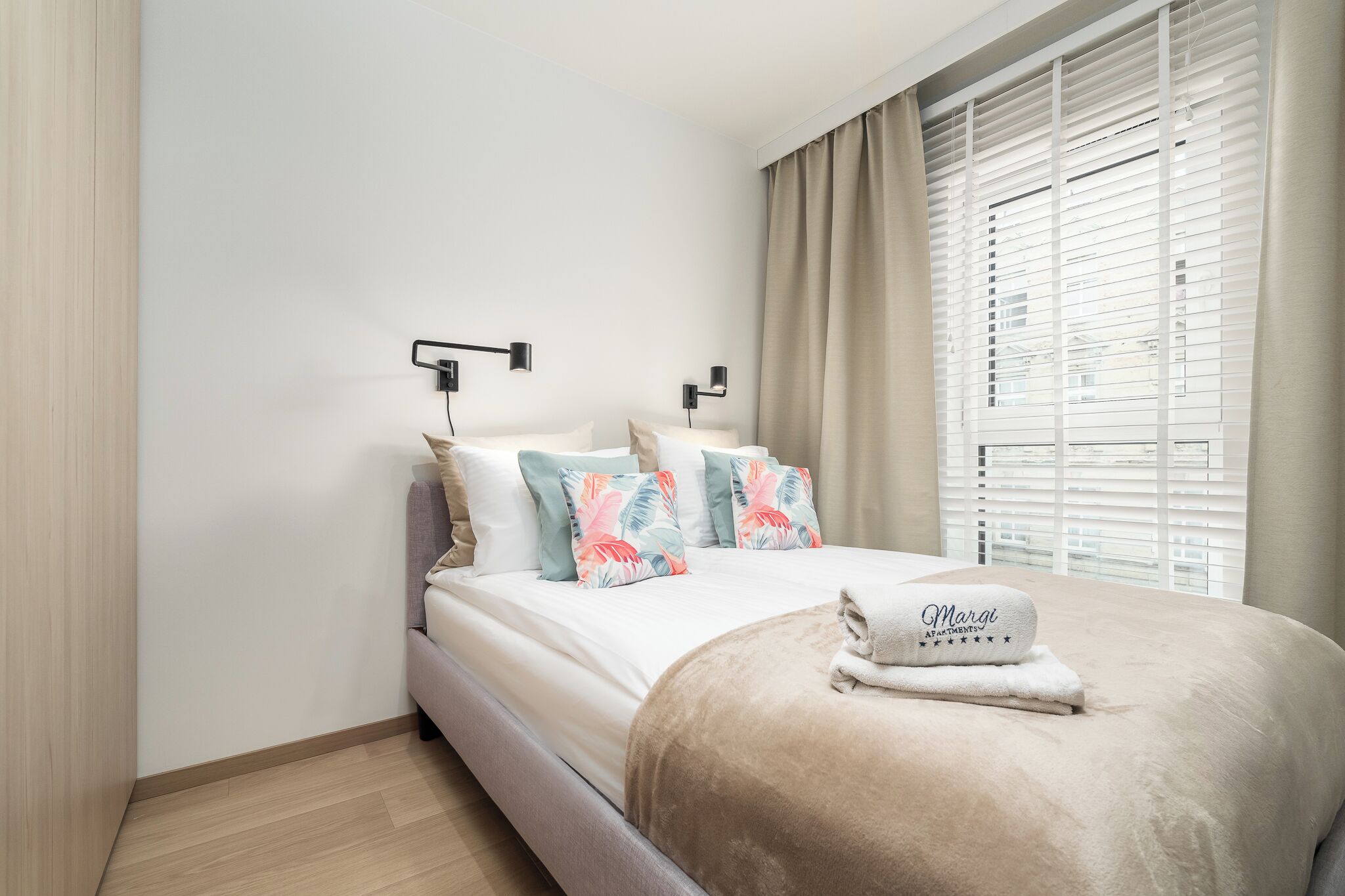 A neatly made bed with a beige blanket and a towel with 'Margo Apartments' text, white bedding, and floral pillows.