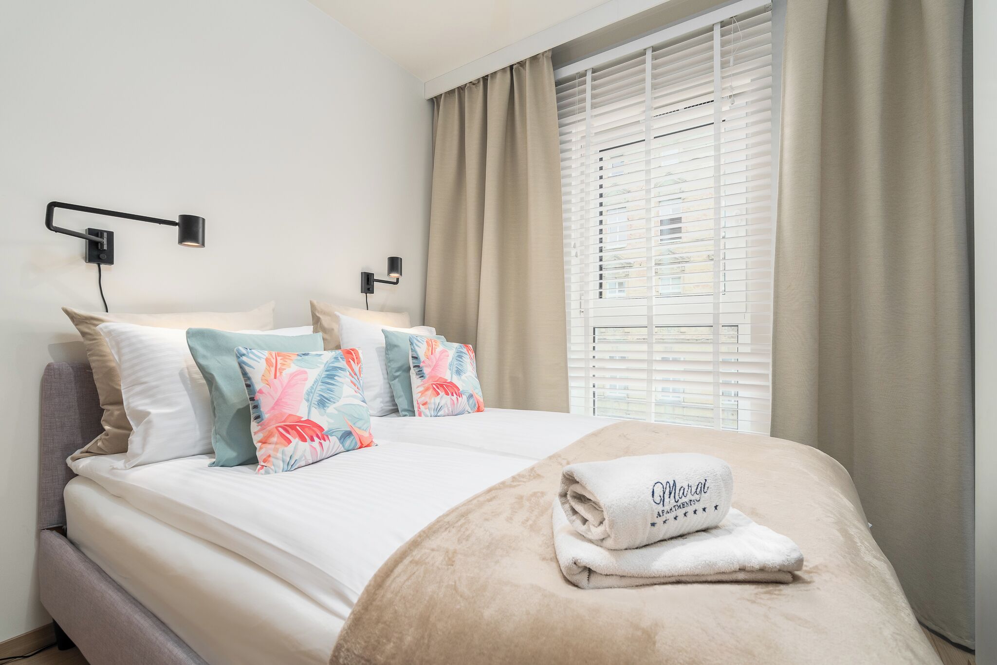 A neatly made bed with a beige blanket and a towel with text, a headboard, and a bedside lamp.