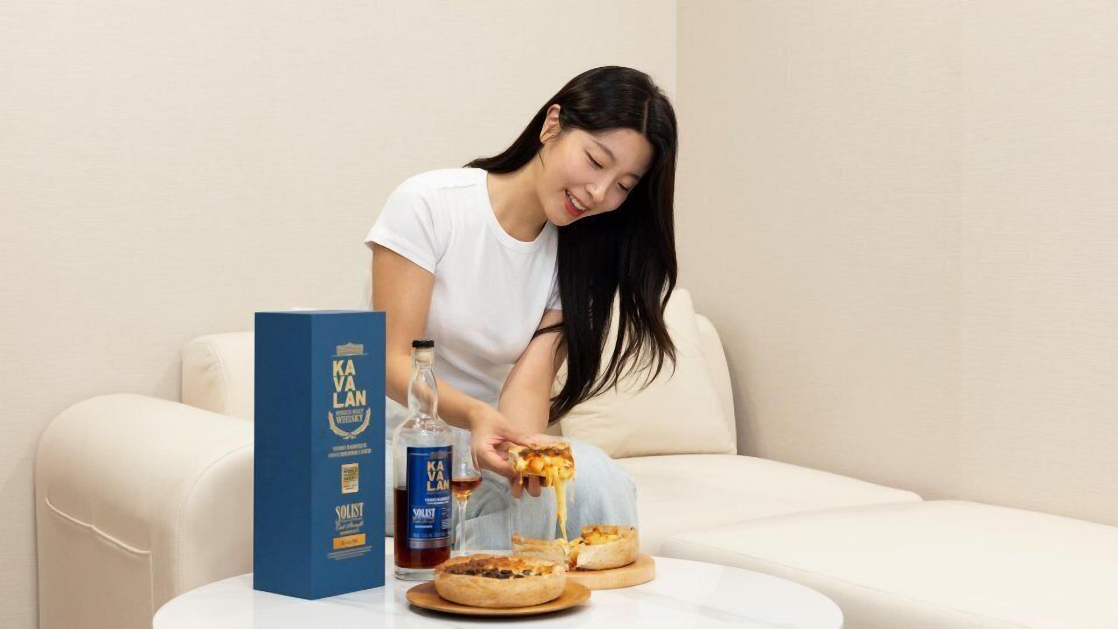 Une personne est assise sur un canapé, avec une boîte de pizza, une bouteille de whisky et une bouteille de vin sur une petite table.