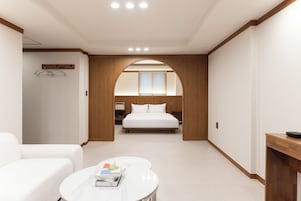 Una habitación de hotel moderna con una cama, un sofá blanco, una mesita con libros y un arco de madera que da acceso al dormitorio.