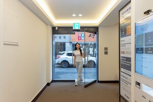 Una persona de pie en el vestíbulo de un hotel, con un vidrio que da a la calle, una máquina expendedora y un letrero con texto coreano.