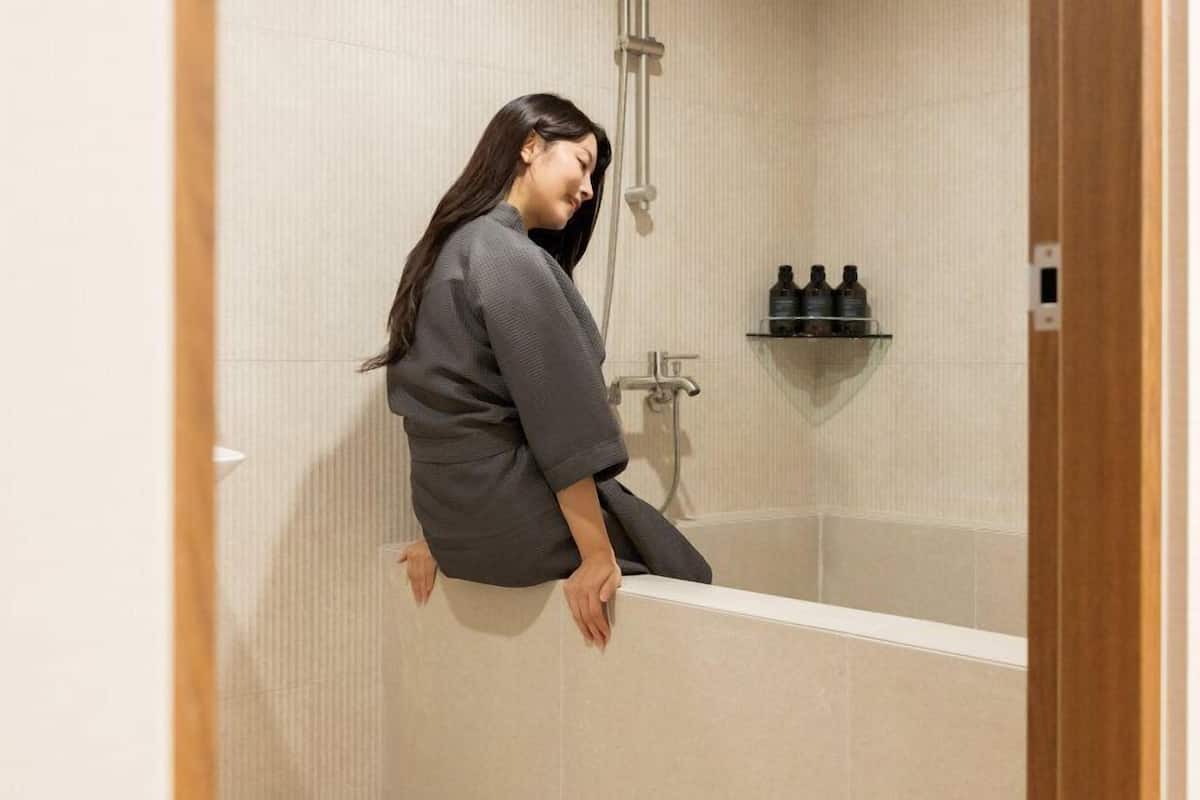 Une personne est assise sur le bord d’une baignoire dans une salle de bain dont les murs sont recouverts de carreaux et qui comporte une étagère avec des bouteilles.
