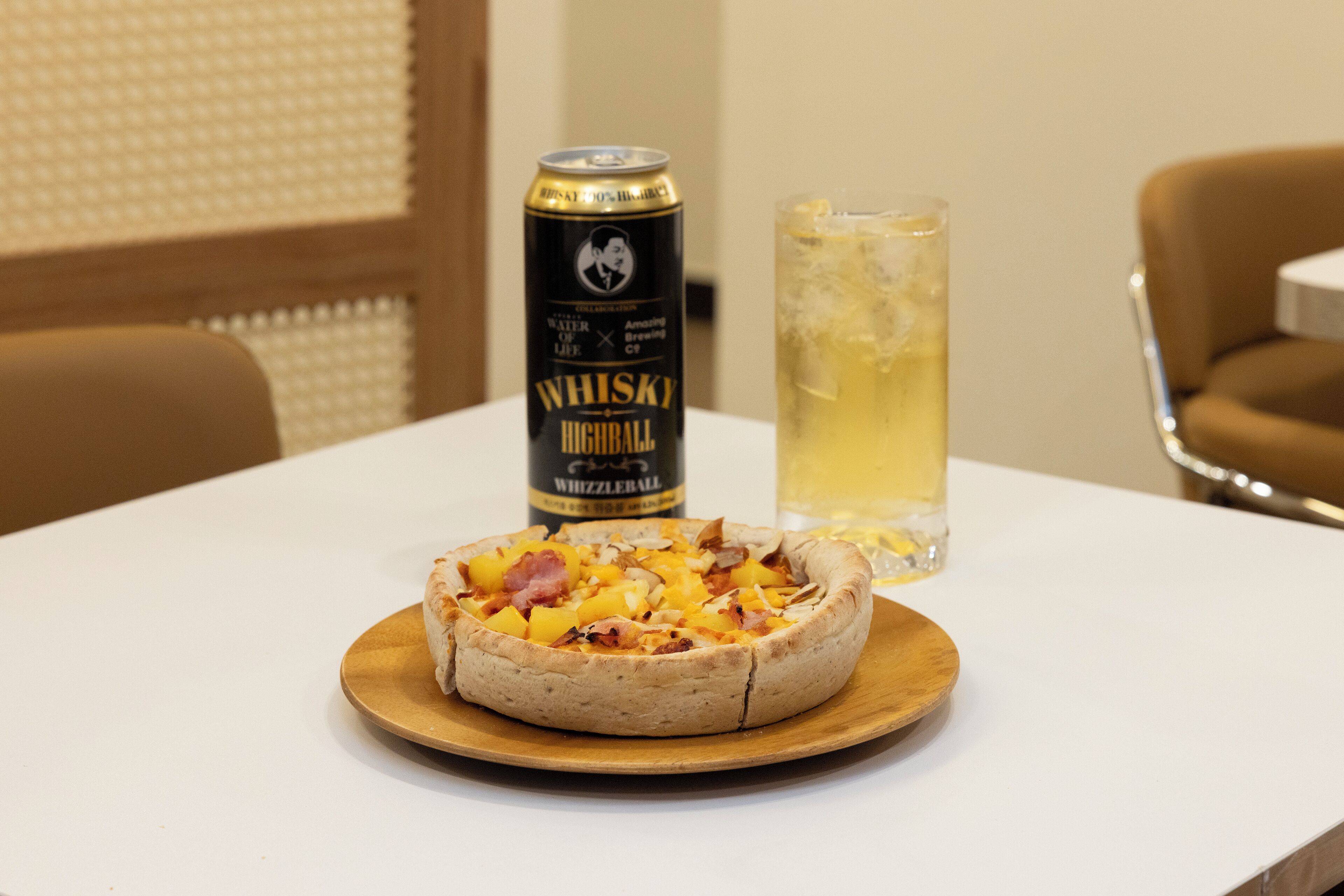 Una pizza sobre una tabla de madera en una mesa blanca. También hay una lata de whisky y un vaso alto de cerveza sobre la mesa.
