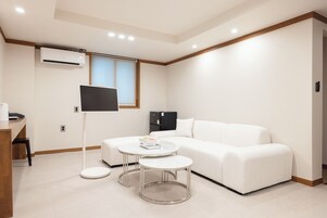 Quarto de hotel moderno com sofá branco, duas mesas redondas, televisão de tela plana, ar condicionado e janela com persianas.