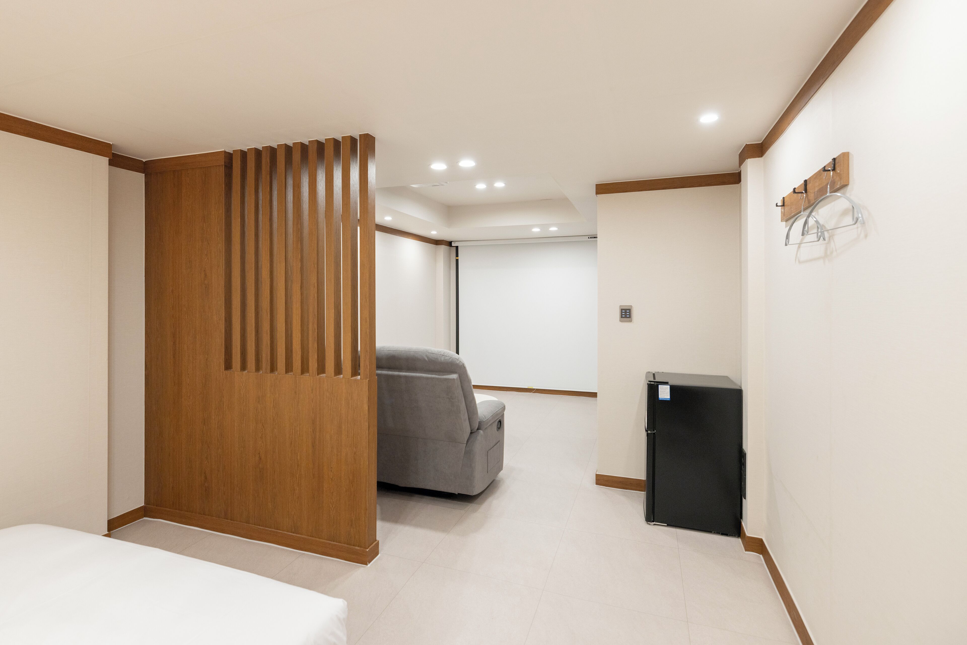 A modern hotel room with a wooden partition, a gray sofa, a small black refrigerator, and a white bed.