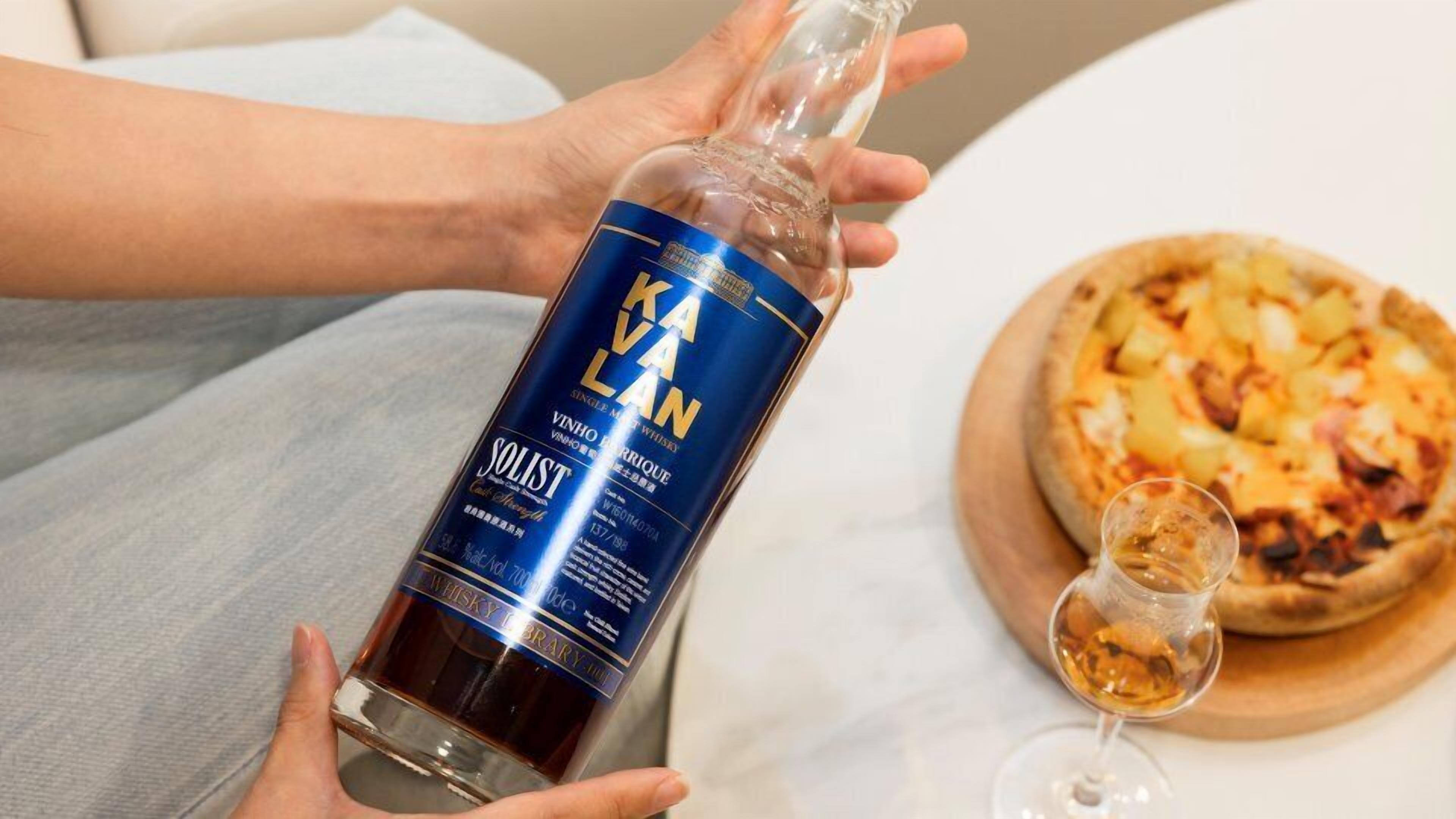 A person holding a bottle of Solist wine, with a pizza and a glass of wine on a table.