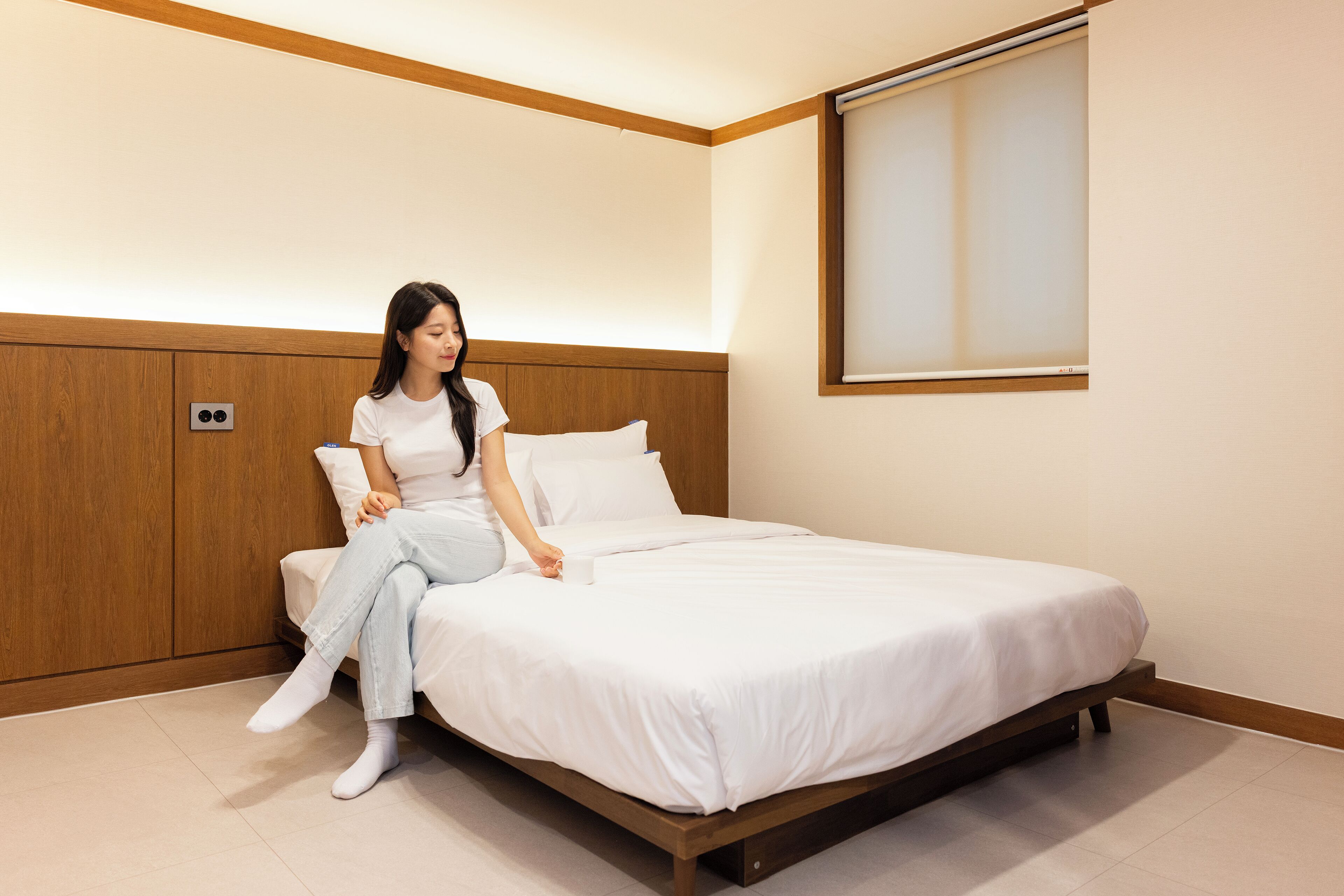 A neatly made bed with white linens in a room with wooden paneling and a window with blinds.
