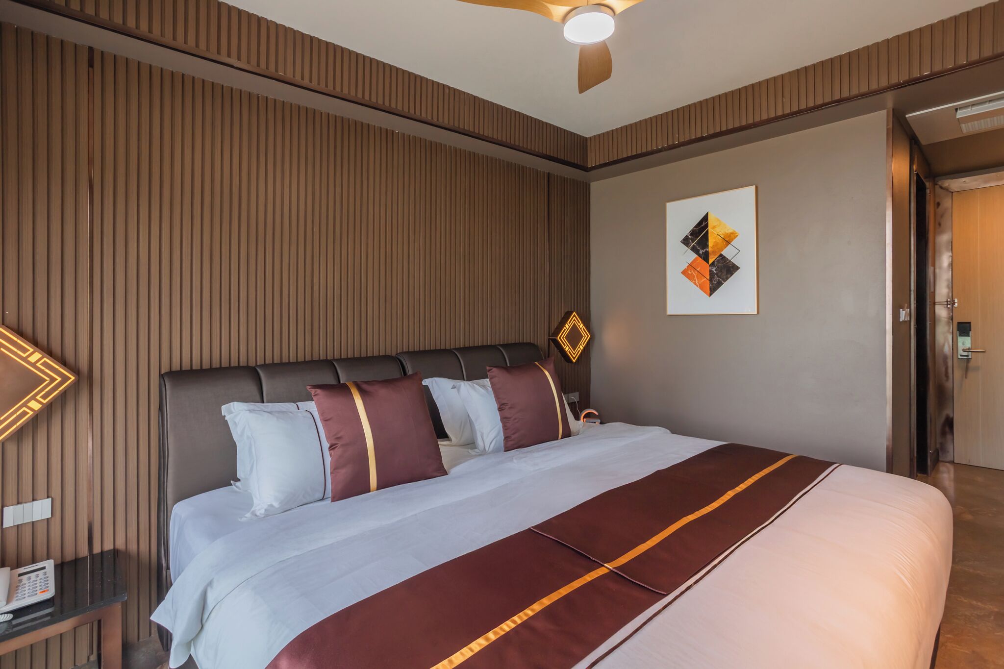 A hotel room with a large bed, a wooden panel wall, a ceiling fan, a bedside table with a phone, and a door to the right.