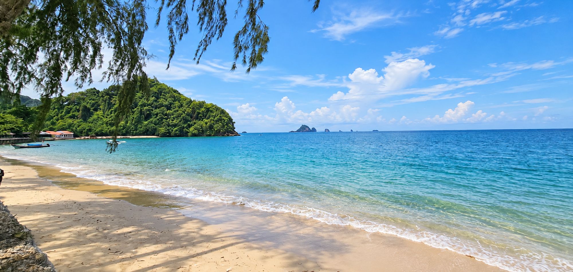 A beach with clear turquoise water, sandy shore, and a forested hill in the background.