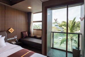 Habitación de hotel con una cama grande, una mesita de noche con teléfono, un área de descanso con un banco y cojines, y un balcón con vistas a palmeras y edificios.