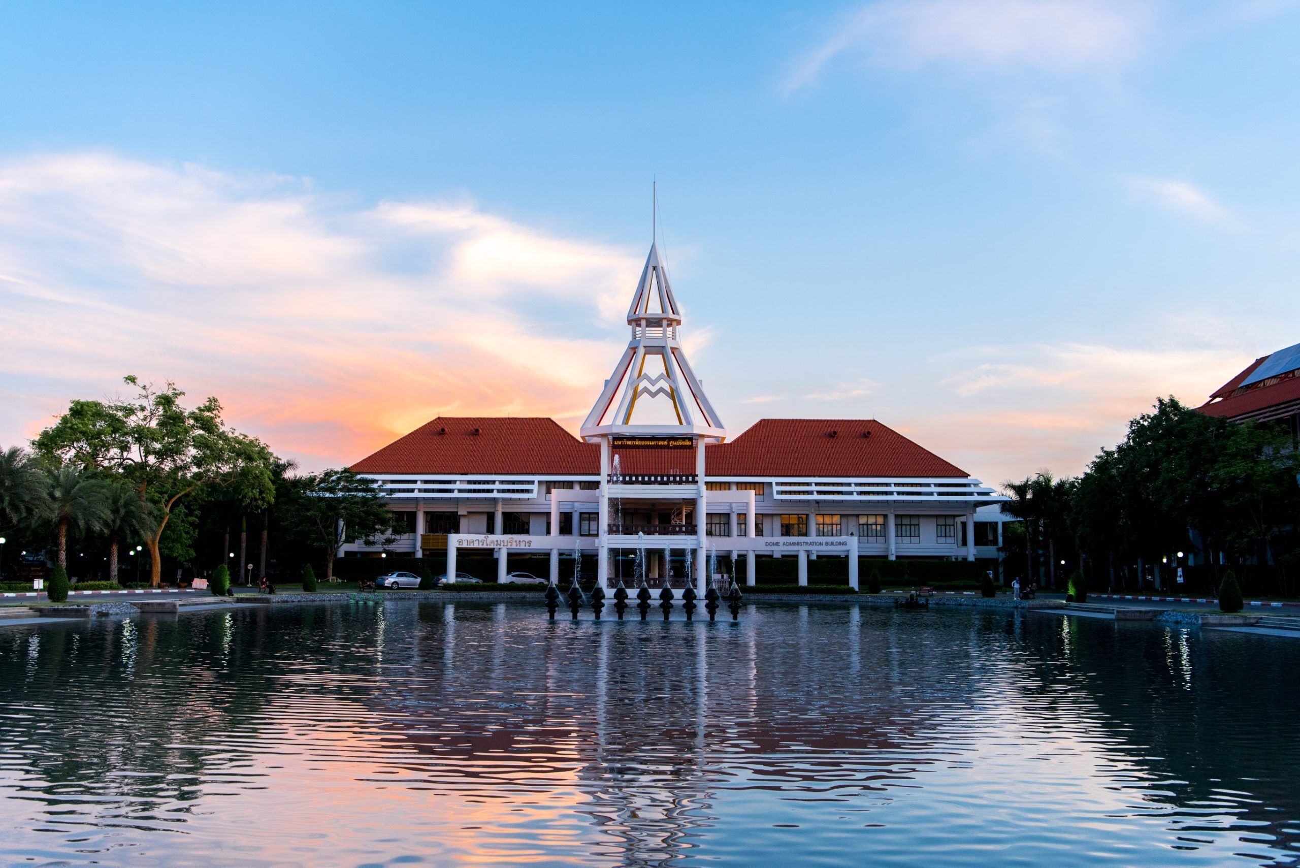 A building with a central tower, a water body in front, and a red-roofed structure to the right.