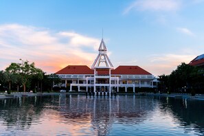 A building with a central tower, a water body in front, and a red-roofed structure to the right. - THE MOMENT (Khlong Luang)