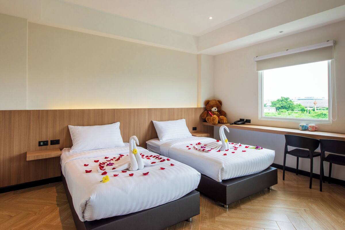 Habitación de hotel con dos camas, un escritorio, una silla, un osito de peluche y una ventana con vistas.